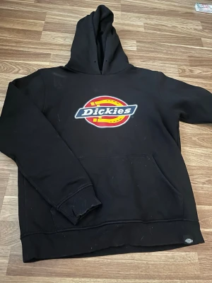 Svart Dickies hoodie med logga - Svart hoodie från Dickies med stor färgglad logga på bröstet. Klassisk modell med huva och känguruficka framtill. Mjuk bomullskänsla och ribbade muddar vid ärmslut och nederkant. Perfekt för en avslappnad streetwear-look.
