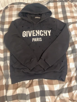 Svart hoodie från Givenchy Paris - Svart hoodie från Givenchy Paris med vit logga framtill. Hoodien har huva med dragsko och känguruficka. Distinkt streetwear-look med slitna detaljer i tyget. Perfekt för dig som gillar exklusiva märken och vill sticka ut.