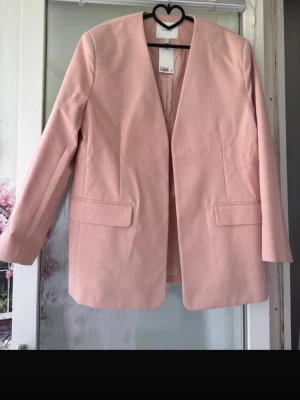 Ljusrosa kavaj från H&M Edition - Stilren ljusrosa kavaj från H&M Edition i rak modell med långa ärmar och utan knäppning. Kavajen har två fickor med lock framtill och är tillverkad i ett slätt tyg som ger en elegant look. Perfekt för att lyfta din outfit med en soft pastellfärg.