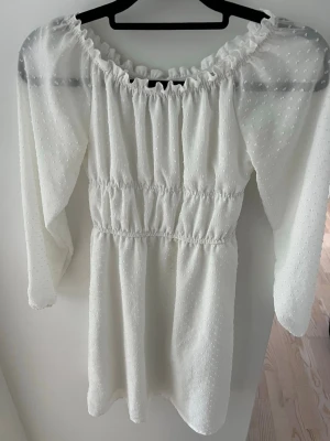 Vit kort sommarklänning med tunn ärm - Supersöt vit kort klänning med smock i midjan och puffiga långärmar. Klänningen har en offshoulder-ringning och är gjord i ett lätt, prickigt vitt tyg som ger en romantisk känsla. Perfekt för sommarens alla tillfällen! Divided i strl xxs