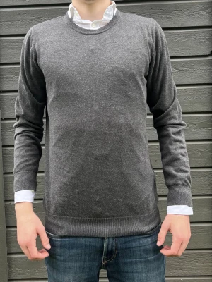 En riktig fredag stickad tröja från dressman!  - Mörkgrå stickad tröja från Dressmann med rund halsringning och långa ärmar. Tröjan har ribbade muddar vid ärmslut och nederkant, vilket ger en snygg och klassisk look. Perfekt att bära över en skjorta för en stilren outfit. Tröjan är riktig fet till en utekväll och ger fräscha vibbar. Ifall du undrar något ställ gärna en fråga!