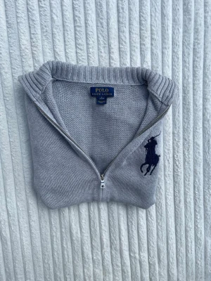 Grå ziptröja från Polo Ralph Lauren - Snygg grå ziptröja från Polo Ralph Lauren med stor mörkblå logga på bröstet och siffran 3 på ärmen. Tröjan har ribbade muddar, två fickor fram och hög krage. Perfekt för dig som gillar klassisk och sportig stil.