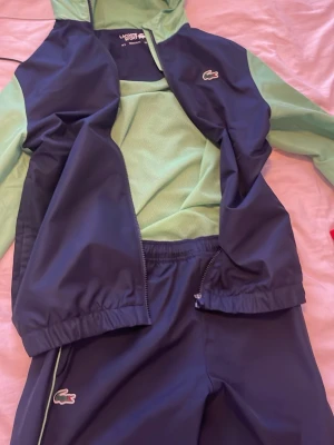 Lacoste dress - Säljer min Lacoste dress i S storlek. Koftan är blågrön med luva. Byxorna är blåa och raka och har en dragkedja längst ner som öppnar upp byxorna. Perfekt för blåsigt väder