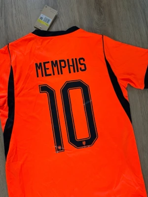 Nederländerna fotbollströja Memphis - Snygg orange fotbollströja från Nike med svarta detaljer, officiell Holland landslagströja med Memphis Depay och nummer 10 på ryggen. Kort ärm, svart krage och landslagets emblem på bröstet. Perfekt nu inför VM2026. 
