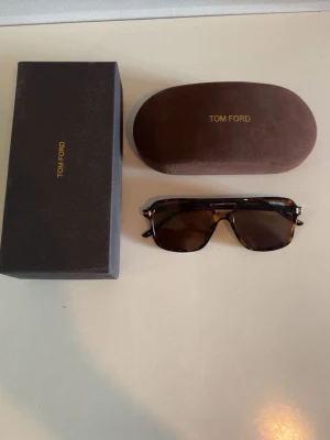 Tom Ford bruna solglasögon - Snygga solglasögon från Tom Ford med brunmelerad båge och mörka rektangulära glas. Klassisk och stilren design med diskret Tom Ford-logga på sidan. Kommer med originalfodral och putsduk.