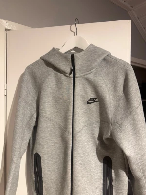 Grå Nike hoodie med dragkedja - Snygg grå hoodie från JD Sports med svart dragkedja framtill och två fickor med svarta detaljer. Klassisk Nike-logga på bröstet och huva för extra komfort. Tillverkad i mjukt material som passar perfekt till en avslappnad stil.