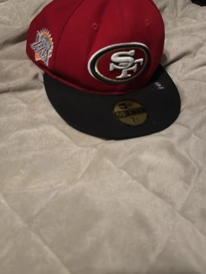Röd 49ers keps från New Era - Säljer en röd och svart snapback-keps från New Era med stort broderat San Francisco 49ers-logo framtill och Super Bowl XXIX-märke på sidan. Klassisk flat brim och sticker kvar på skärmen. Perfekt för dig som gillar sportig streetstyle.