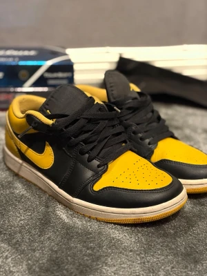 Nike Air Jordan 1 Low svart/gul - Snygga Nike Air Jordan 1 Low sneakers i svart och gult läder med klassisk Air Jordan-logga broderad på hälen. Skorna har gula sulor och svarta skosnören. Perfekta för dig som vill sticka ut med en ikonisk och sportig look.