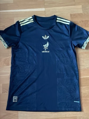 Adidas Mexiko svart sporttröja - Snygg svart sporttröja från Adidas med guldfärgade detaljer och Mexiko-tryck på bröstet. Tröjan har korta ärmar, rund hals och diskreta mönster i tyget. Tre guldiga ränder på axlarna och dekorativa muddar vid ärmsluten. Perfekt för träning eller match.