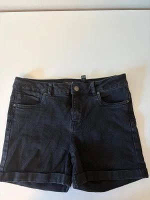 Svarta denimshorts från Vero Moda - Snygga svarta denimshorts från Vero Moda med klassisk femficksmodell och uppvikta benslut. Shortsen har knapp och dragkedja framtill samt bälteshällor. Perfekta för en avslappnad och trendig sommarstil.