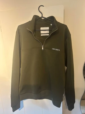 Olivgrön halfzip hoodie Les Deux - Snygg olivgrön hoodie från Les Deux med halv dragkedja och vit logga på bröstet. Klassisk passform med ribbade muddar och hög krage. Perfekt för en avslappnad men stilren look.
