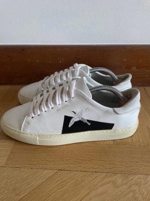 Vita sneakers från Axel Arigato - Snygga vita sneakers från Axel Arigato med svart detalj och broderad fågel på sidan. Skorna har klassisk låg profil, vita skosnören och beige sula. Tillverkade i läder för en clean och stilren look som passar till det mesta.