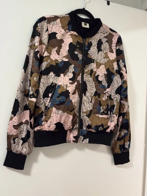 Mönstrad bomberjacka från PEP - Snygg bomberjacka från PEP med ett unikt abstrakt mönster i beige, rosa, svart, blått och brunt. Jackan har svarta muddar, dragkedja framtill och två fickor. Perfekt för dig som vill sticka ut med en cool och färgstark look.