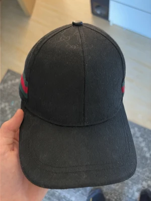 Svart keps från Gucci med ränder - Snygg svart keps från Gucci med diskret GG-mönster i tyget och klassiska gröna och röda ränder på sidan. Kepsen har böjd skärm och justerbar passform. Perfekt för dig som vill ha en stilren accessoar med lyxig känsla.