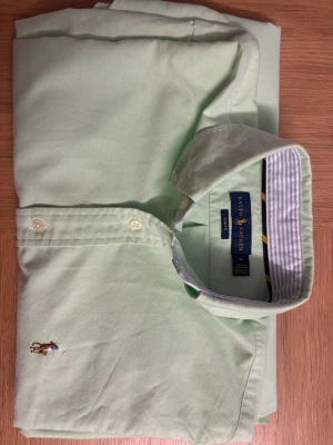 Ralph Lauren skjorta  - Snygg ljusgrön skjorta från Ralph Lauren med klassisk krage och broderad logga på bröstet. Skjortan har knappar framtill och är tillverkad i mjuk bomull. Perfekt för en stilren och avslappnad look.
