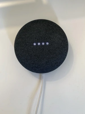 Google Nest Mini - Säljer en Google Nest Mini, en kompakt och rund smart högtalare i svart tygmaterial. Den har fyra LED-lampor på ovansidan och ansluts med vit sladd. Perfekt för att styra musik, smarta hemmet och få svar direkt med rösten.