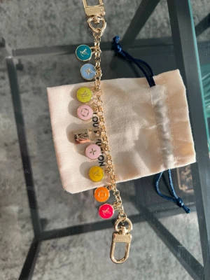 Bag charm  färgglatt charmarmband - Snyggt bag charm med guldfärgad kedja och färgglada berlocker i blått, grönt, rosa, gult, orange och rött. Berlockerna har LV-logga och monogramblommor. Perfekt accessoar för dig som gillar lyxiga detaljer och färg.