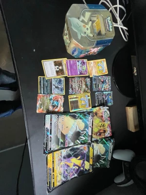 Pokémonkort samling med plåtlåda - Samling Pokémonkort i olika färger och motiv, inklusive flera holografiska och specialkort. Medföljer en plåtlåda med Togepi-design för förvaring av korten. Perfekt för dig som gillar att samla eller spela Pokémon TCG.