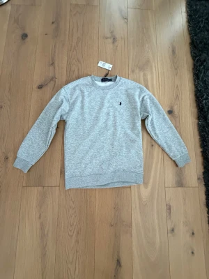 Grå sweatshirt från Polo Ralph Lauren - Klassisk grå sweatshirt från Polo Ralph Lauren med rund halsringning och den ikoniska lilla broderade loggan på bröstet. Tröjan har ribbade muddar vid ärmslut och nederkant. Perfekt till jeans eller joggers för en avslappnad look.