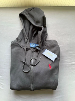 Svart hoodie från Polo Ralph Lauren - Säljer en svart hoodie från Polo Ralph Lauren med dragkedja, huva och dragsnören. Klassisk röd logga broderad på bröstet och två fickor framtill. Tillverkad i mjukt bomullsmaterial som är skönt att ha på sig.