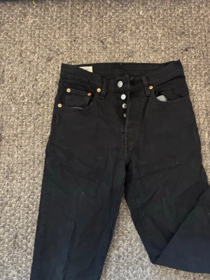 Svarta Levi's 501 jeans med knappar - Säljer ett par klassiska svarta Levi's 501 jeans med knappgylf och raka ben. Jeansen har fem fickor, silverfärgade knappar och detaljer i koppar. Tillverkade i kraftig denim med den ikoniska läderlappen bak i midjan.