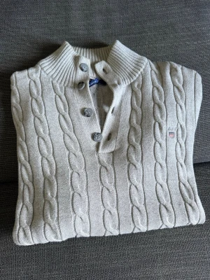 Beige kabelstickad tröja från Gant - Kabelstickad tröja från Gant i beige bomull med ribbad hög krage och fyra knappar framtill. 