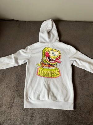 Vit Spongebob hoodie med tryck - Vit hoodie med stor färgglad Spongebob-tryck på ryggen och texten 'IS THE COLOR OF HAPPINESS'. På bröstet står det 'SPONGEBOB' i rosa och gult. Hoodien har huva med dragsko och en klassisk magficka. Perfekt för dig som gillar streetwear och tecknade motiv.