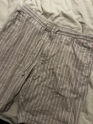 Beige randiga linneshorts från H&M - Säljer ett par beige shorts från H&M i skönt linnematerial. Shortsen har vita vertikala ränder och snörning i midjan för en avslappnad look. Perfekta för varma dagar och har en loose passform som ger extra komfort.