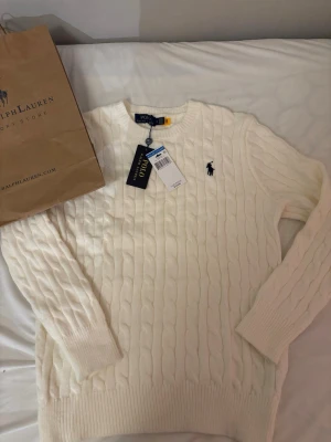 Vit kabelstickad tröja Polo Ralph Lauren - Kabelstickad tröja i krämvit färg från Polo Ralph Lauren. Klassisk rund halsringning, långa ärmar och ribbade muddar. Tröjan har den ikoniska broderade loggan på bröstet och är tillverkad i mjukt material för en stilren look.