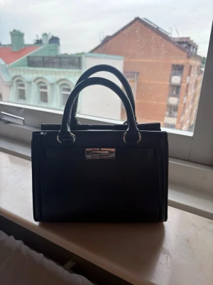 Svart handväska - Snygg svart handväska med dubbla handtag och silverfärgat metallspänne framtill. Väskan har ett rymligt huvudfack och är tillverkad i ett slätt material som ger en stilren look. Perfekt för dig som gillar klassiska accessoarer.