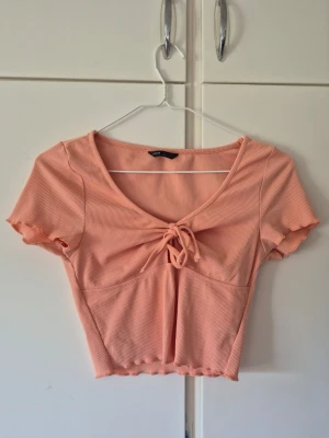 Aprikos ribbad croppad topp med knyt - Söt aprikos croppad topp med ribbad struktur och knyt framtill. Toppen har korta ärmar med vågig kant och en djup v-ringning. Perfekt för varma dagar och enkel att styla till jeans eller kjol. Säljer då den inte används längre