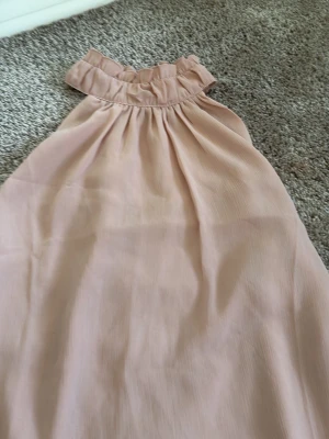 Ljusrosa ärmlös blus från H&M - Supersöt ljusrosa topp från H&M med volangdetalj vid halsen och knappar i nacken. Toppen är ärmlös och har en lös passform i ett mjukt, lätt material som känns skönt mot huden. Perfekt för varma dagar eller när du vill känna dig extra fin.