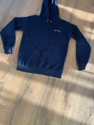 Mörkblå hoodie med text Ma chérie - Mörkblå hoodie med huva och dragsko. På bröstet står det 'Ma chérie' broderat i vitt. Klassisk passform med ribbade muddar vid ärmar och nederkant. Perfekt för en chill och stilren look. Materialet känns mjukt och bekvämt, troligen bomullsblandning.