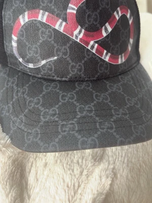  keps med orm och GG-mönster - Säljer en svart keps  med klassiskt GG-mönster och en rödvit orm på framsidan. Kepsen har böjd skärm och är tillverkad i slitstarkt tyg. Perfekt accessoar för dig som vill sticka ut med exklusiv streetstyle. Har inte andvänd på länge 