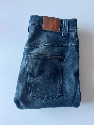 Blå jeans från Nudie Jeans - Säljer ett par blå jeans från Nudie Jeans med klassisk femficksdesign och brunt läderpatch bak. Jeansen har en slim fit och är tillverkade i väldigt rå denim. Storleken är 30/34 men sitter lite kortare än så. Skriv såklart vid frågor eller funderingar🙌