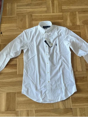Vit skjorta från Polo Ralph Lauren - Klassisk vit skjorta från Polo Ralph Lauren med broderad marinblå logga på bröstet. Skjortan har lång ärm, knappar framtill och button-down krage. Tillverkad i mjuk bomull för en clean och stilren look.