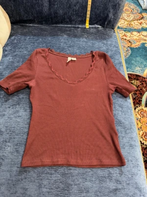 Brun ribbad topp med spets från H&M - Säljer en brun ribbad topp från H&M Divided i storlek S. Toppen har korta ärmar, v-ringning med spetsdetalj och sitter snyggt på kroppen. Perfekt att styla med jeans eller kjol för en trendig look.