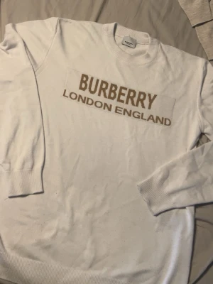 Vit Burberry tröja med logga - Snygg vit tröja från Burberry med stor beige logga och texten 'London England' framtill. Tröjan har rund halsringning, långa ärmar och ribbade muddar. Perfekt för dig som gillar stilrena och exklusiva plagg.