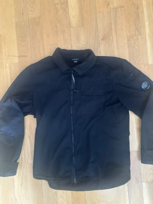 Svart overshirt från C.P. Company - Stilren svart overshirt från C.P. Company med dragkedja framtill, klassisk krage och bröstficka. Ärmen har den ikoniska linsen på vänster sida. Tillverkad i ett lätt och slitstarkt material som passar perfekt till lager-på-lager-stil. Inte använd så mycket och perfekt skick