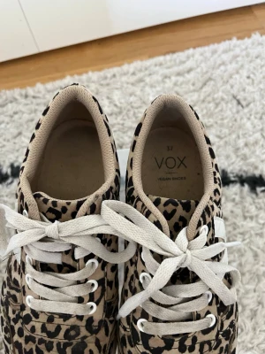 Leopardmönstrade sneakers från VOX - Säljer ett par snygga sneakers från VOX med leopardmönster i beige och svart. Skorna har vita snören och en vit sula. Tillverkade i veganskt material och har en klassisk, chunky form som passar perfekt till streetwear.