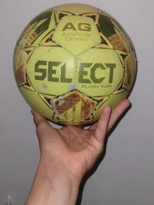 Gul SELECT Flash Turf fotboll - Säljer en gul SELECT Flash Turf fotboll, designad för spel på konstgräs. Bollen har svart och orange grafiskt mönster och texten 'Artificial Grass' tryckt på en av panelerna. Perfekt för dig som älskar fotboll och vill ha en boll för träning eller match. Pris kan diskuteras vid snabb affär!! 🔥🔥🔥 