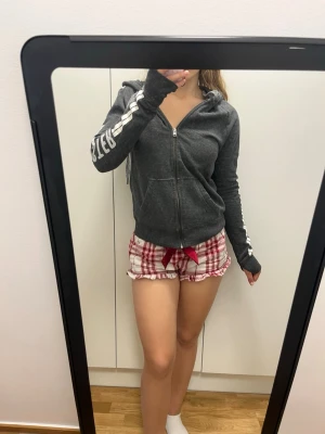 Mörkgrå zip hoodie från Hollister - Mörkgrå hoodie med dragkedja från Hollister. Har vita ränder längs ärmarna och Hollister-tryck på ena ärmen. Justerbar huva med snören och två fickor framtill. Tillverkad i mjukt sweatshirtmaterial, perfekt för en avslappnad stil.