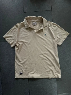 Beige kortärmad t-shirt från Morris - Snygg beige t-shirt från Morris med klassisk krage och liten broderad logga på bröstet. Tillverkad i mjuk bomull och har en avslappnad passform. Perfekt för en clean och enkel stil.