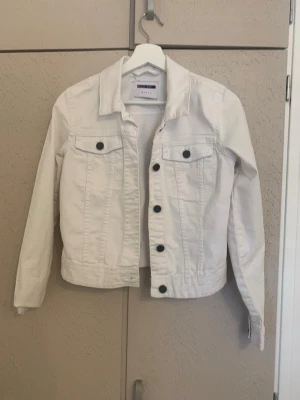 Vit jeansjacka från Noisy May - Snygg vit jeansjacka från Noisy May med klassisk krage, svarta knappar och två bröstfickor med lock. Jackan har en rak passform och långa ärmar, perfekt att slänga över en t-shirt för en clean look.