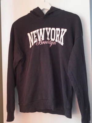 Mörkblå hoodie med New York-tryck - Svart hoodie med stor vit text 'NEW YORK' och mindre text 'Brooklyn' på bröstet. Klassisk modell med huva och ribbade muddar vid ärmar och nederkant. Perfekt för en avslappnad streetwear-look. Materialet känns mjukt och bekvämt.