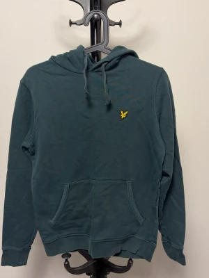 Lyle & Scott - Snygg mörkgrön hoodie från Lyle & Scott med klassisk gul logga på bröstet. Hoodien har dragsko i huvan och en stor känguruficka framtill. Tillverkad i mjuk bomullsmix, perfekt för en avslappnad stil.