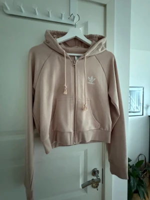 Beige hoodie från Adidas med dragkedja - Snygg beige hoodie från Adidas med dragkedja framtill, två fickor och justerbar huva med snören. Adidas-logga broderad i vitt på bröstet. Tillverkad i mjukt bomullsmaterial som känns skönt mot huden. Perfekt för en avslappnad och sportig stil.