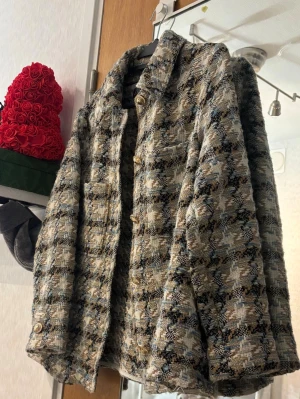 Rutigt tweed cardiganjacka - Snygg cardiganjacka i rutigt tweedmönster med beige, svart, blå och vita toner. Jackan har dekorativa guldknappar framtill och två fickor. Klassisk krage och boxig passform ger en trendig look. Perfekt för lager på lager och streetstyle.