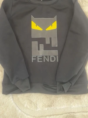 Svart Fendi hoodie med grafiskt tryck - Cool svart hoodie från Fendi med ett stort grafiskt tryck framtill som har gula ögon och Fendi-loggan under. Tröjan har långa ärmar, ribbade muddar och är gjord i mjukt material. Perfekt för dig som gillar streetwear och vill sticka ut.