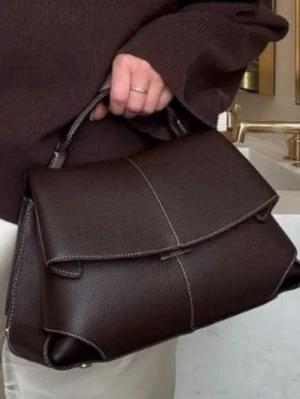 Polene Mörkbrun handväska i läderlook - Stilren mörkbrun handväska från Polene i läderimitation med elegant lock och kontrastsömmar. Väskan har både handtag och avtagbar axelrem, vilket gör den flexibel att bära. Klassisk och rymlig modell med guldfärgade detaljer.